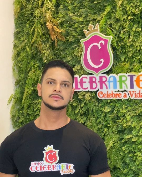Integrante da equipe do Espaço Celebrarte Festas, com a camiseta da casa, na parede de logo do espaço.